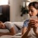 young woman is secretly sending text messages smart phone cheating her boyfriend who is sleeping bed